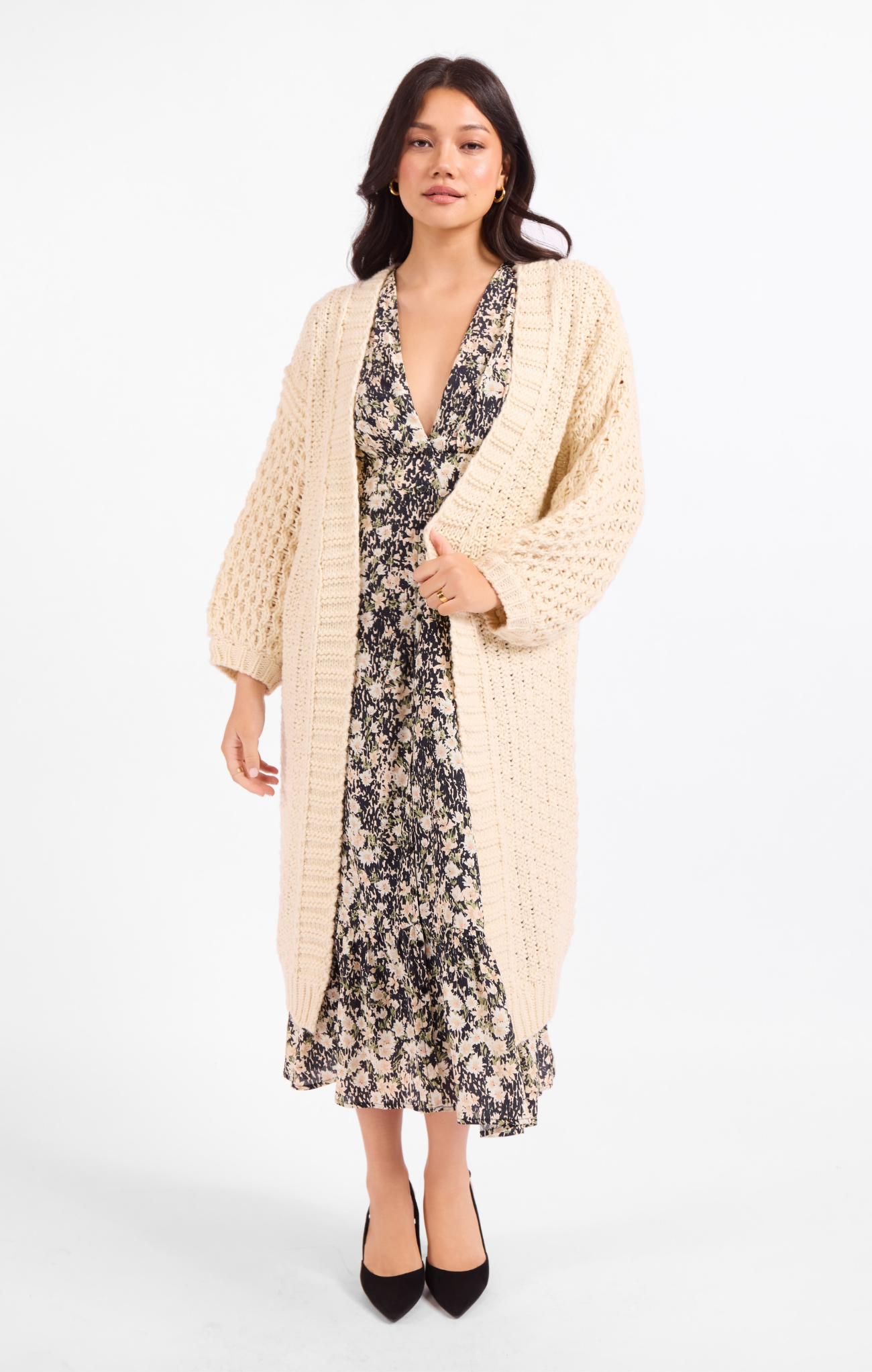 A woman modeling a long cream cable-knit cardigan over a black and beige floral midi dress, paired with black pointed-toe heels, against a plain white studio background.