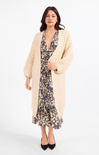 A woman modeling a long cream cable-knit cardigan over a black and beige floral midi dress, paired with black pointed-toe heels, against a plain white studio background.