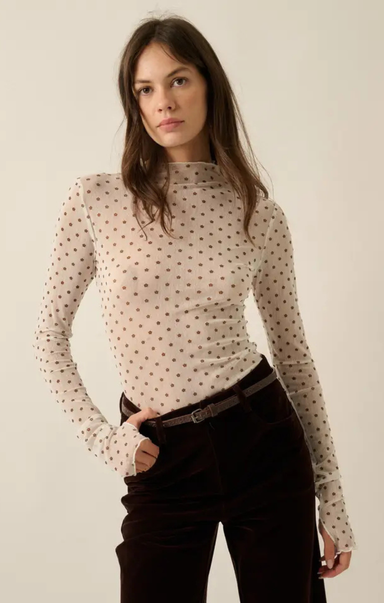 A woman modeling a fitted, long-sleeve mock-neck top in a warm off-white or cream color with a small brown polka dot pattern. She is styled with dark brown corduroy pants and a dark belt, set against a plain, warm off-white studio background.