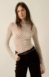 A woman modeling a fitted, long-sleeve mock-neck top in a warm off-white or cream color with a small brown polka dot pattern. She is styled with dark brown corduroy pants and a dark belt, set against a plain, warm off-white studio background.