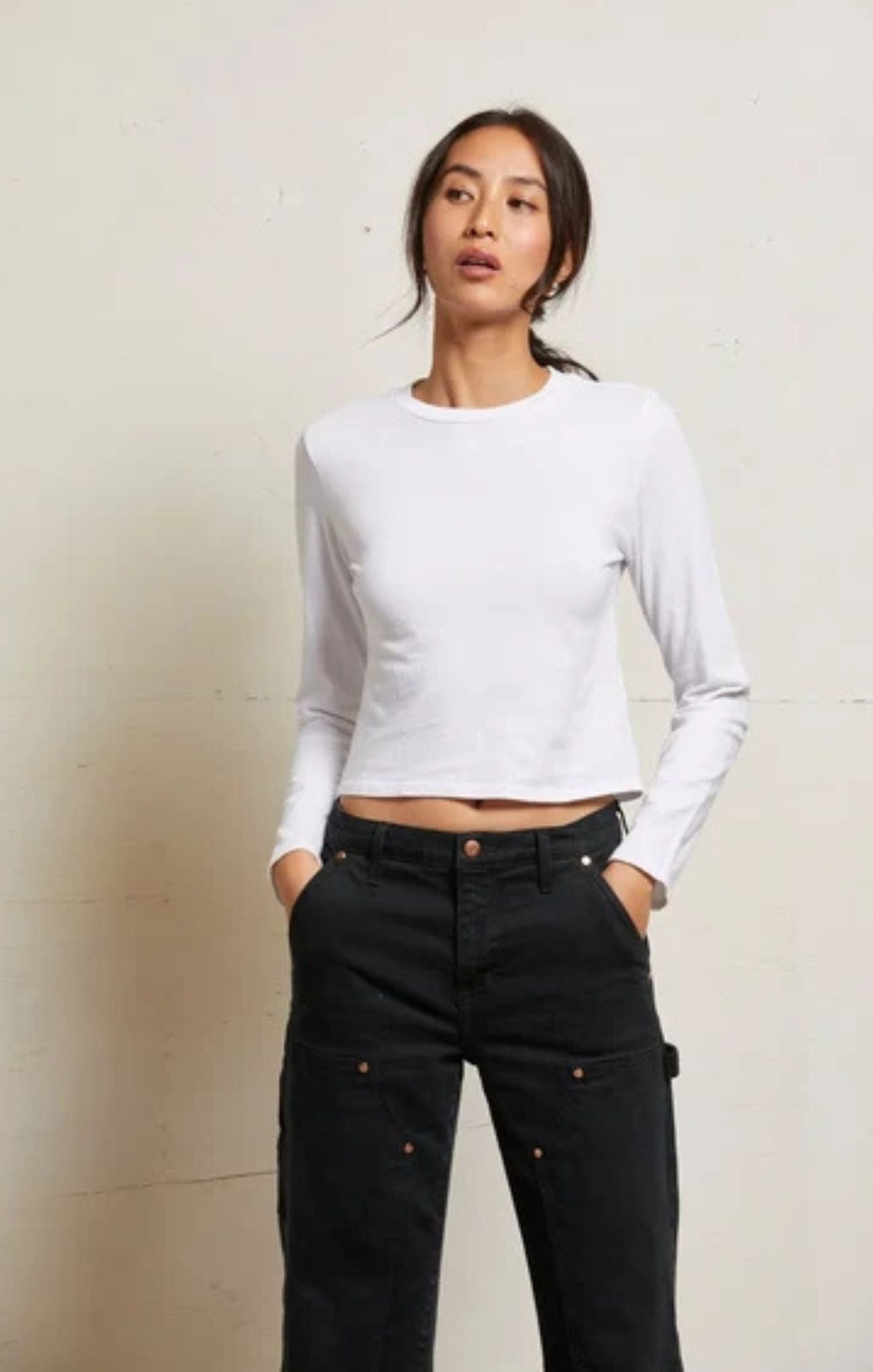 A woman modeling a white long-sleeve crop top paired with black cargo pants, standing against a light beige textured wall.
