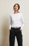 A woman modeling a white long-sleeve crop top paired with black cargo pants, standing against a light beige textured wall.