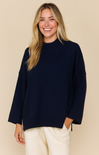 Woman wearing a navy sweater and white pants against a beige background