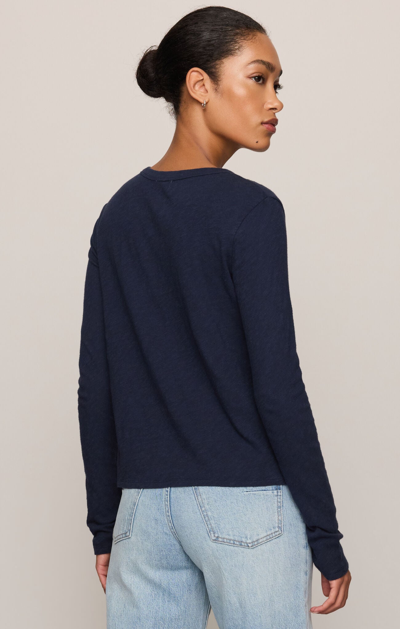 A woman shown from the back wearing a dark navy blue long-sleeve knit sweater and light wash blue jeans, set against a plain light beige studio background.