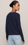 A woman shown from the back wearing a dark navy blue long-sleeve knit sweater and light wash blue jeans, set against a plain light beige studio background.
