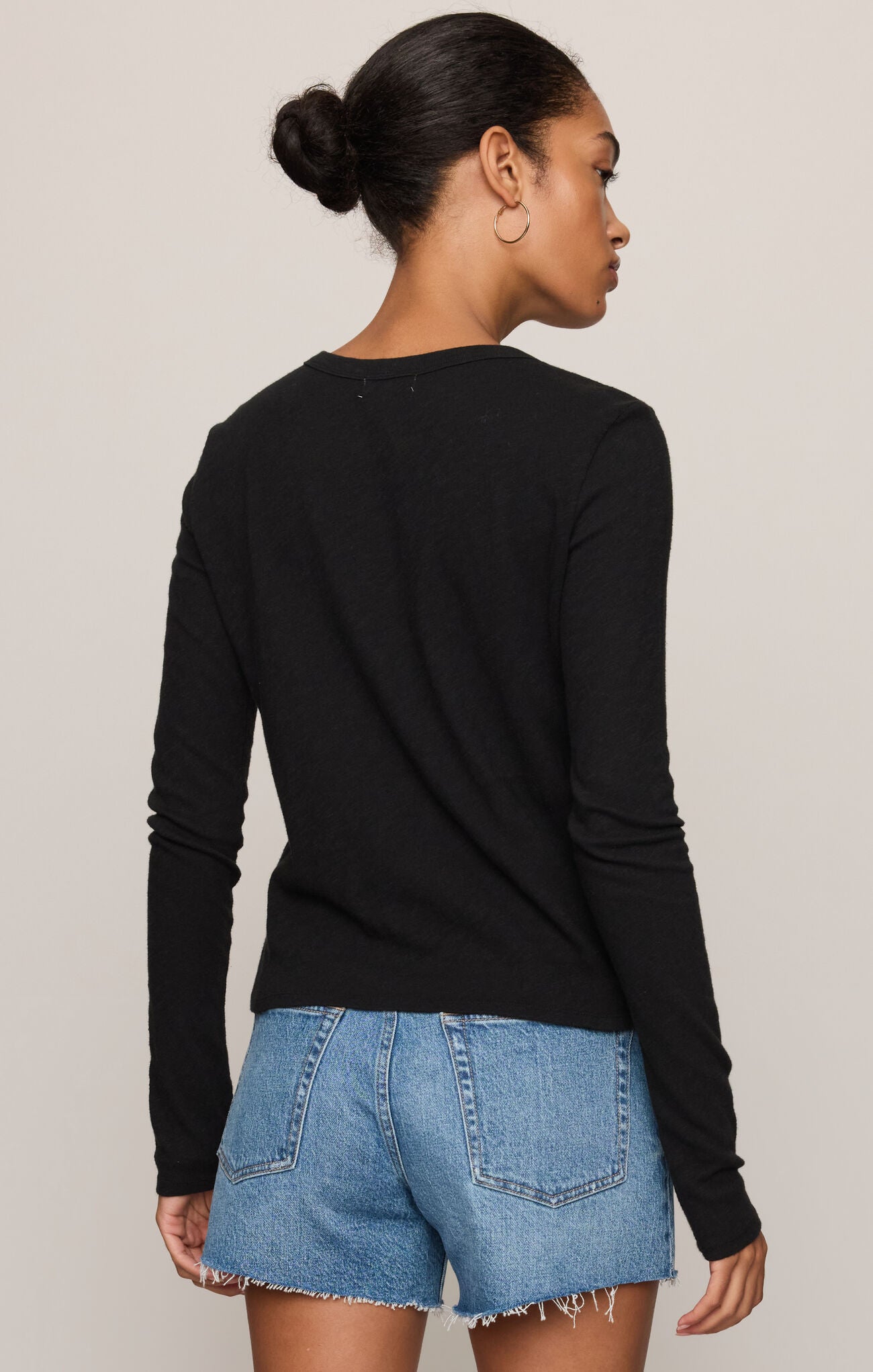 A woman shown from the back wearing a black long-sleeve crewneck top and light blue denim shorts against a plain light gray studio background.
