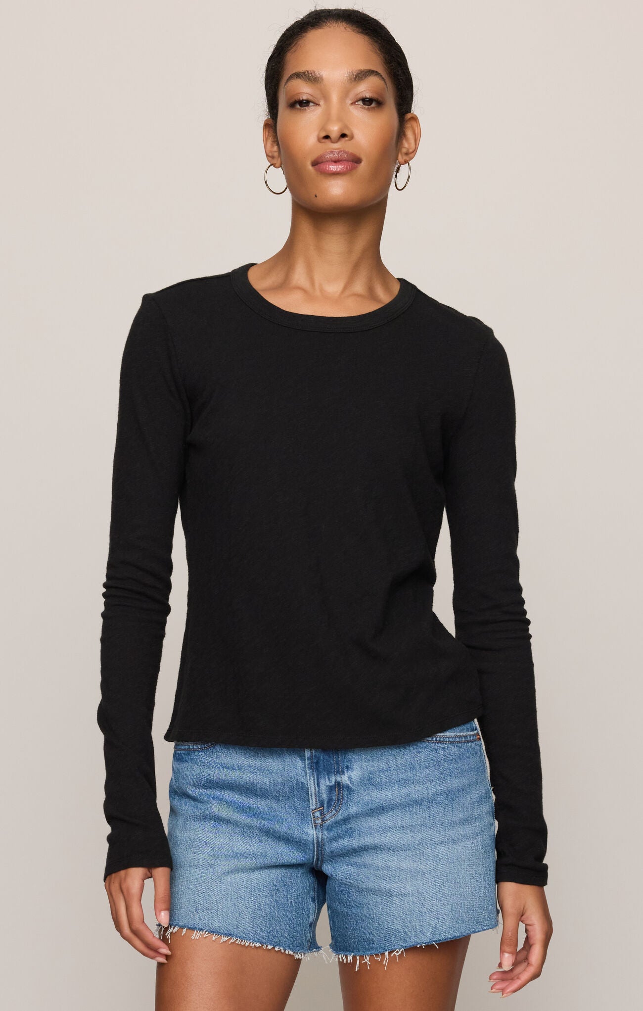 A woman modeling a black long-sleeve crewneck top paired with light blue denim shorts against a solid light beige background.