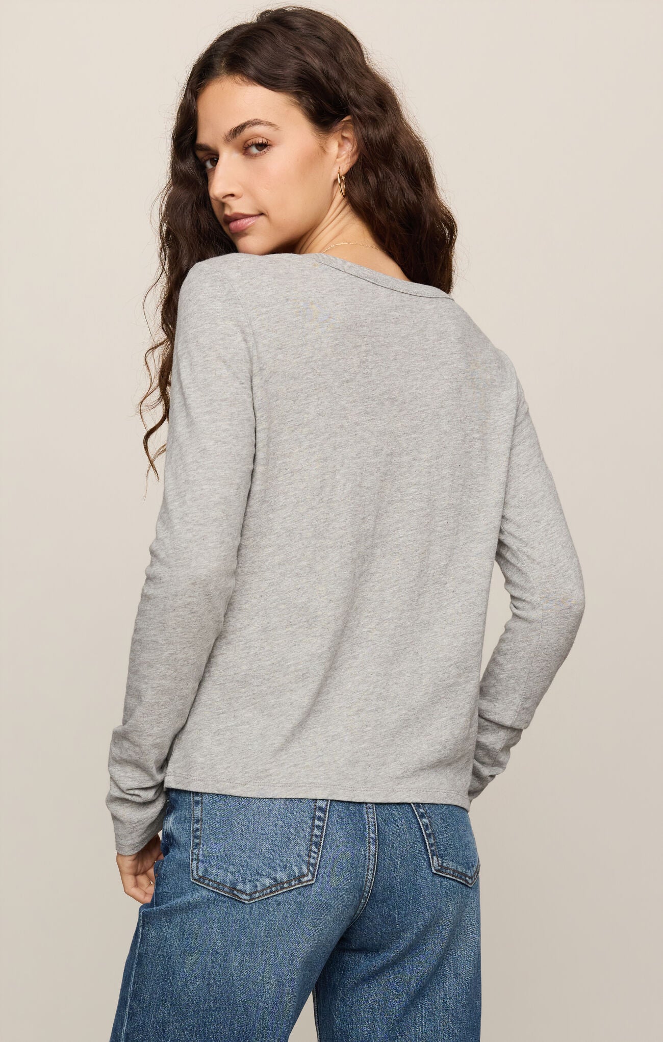 A woman shown from the back wearing a light gray long-sleeve top and blue jeans against a plain beige studio background.