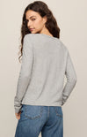 A woman shown from the back wearing a light gray long-sleeve top and blue jeans against a plain beige studio background.