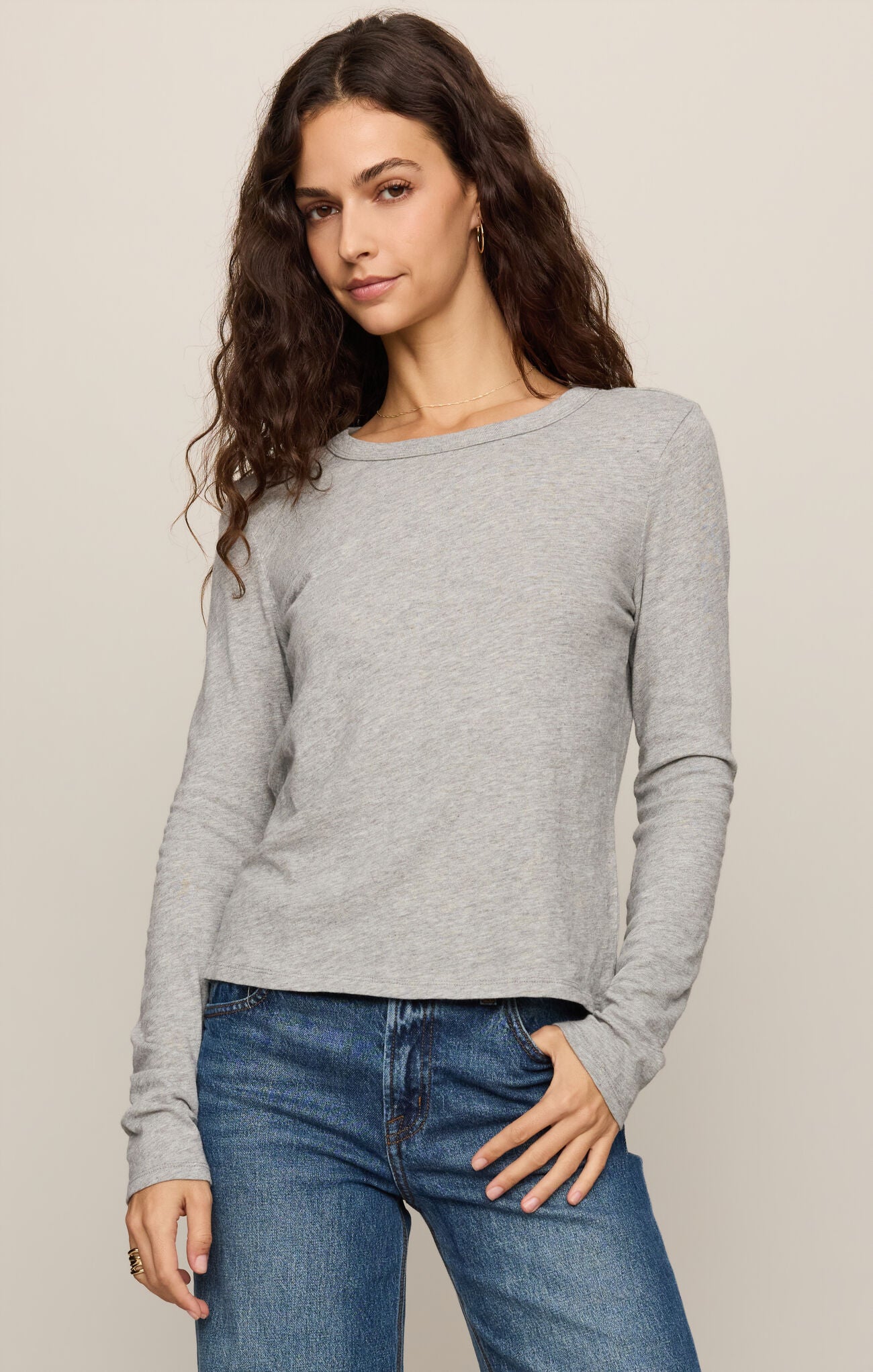 A woman modeling a light gray long-sleeve knit top with a wide boat neckline, paired with blue jeans, against a plain beige studio background.