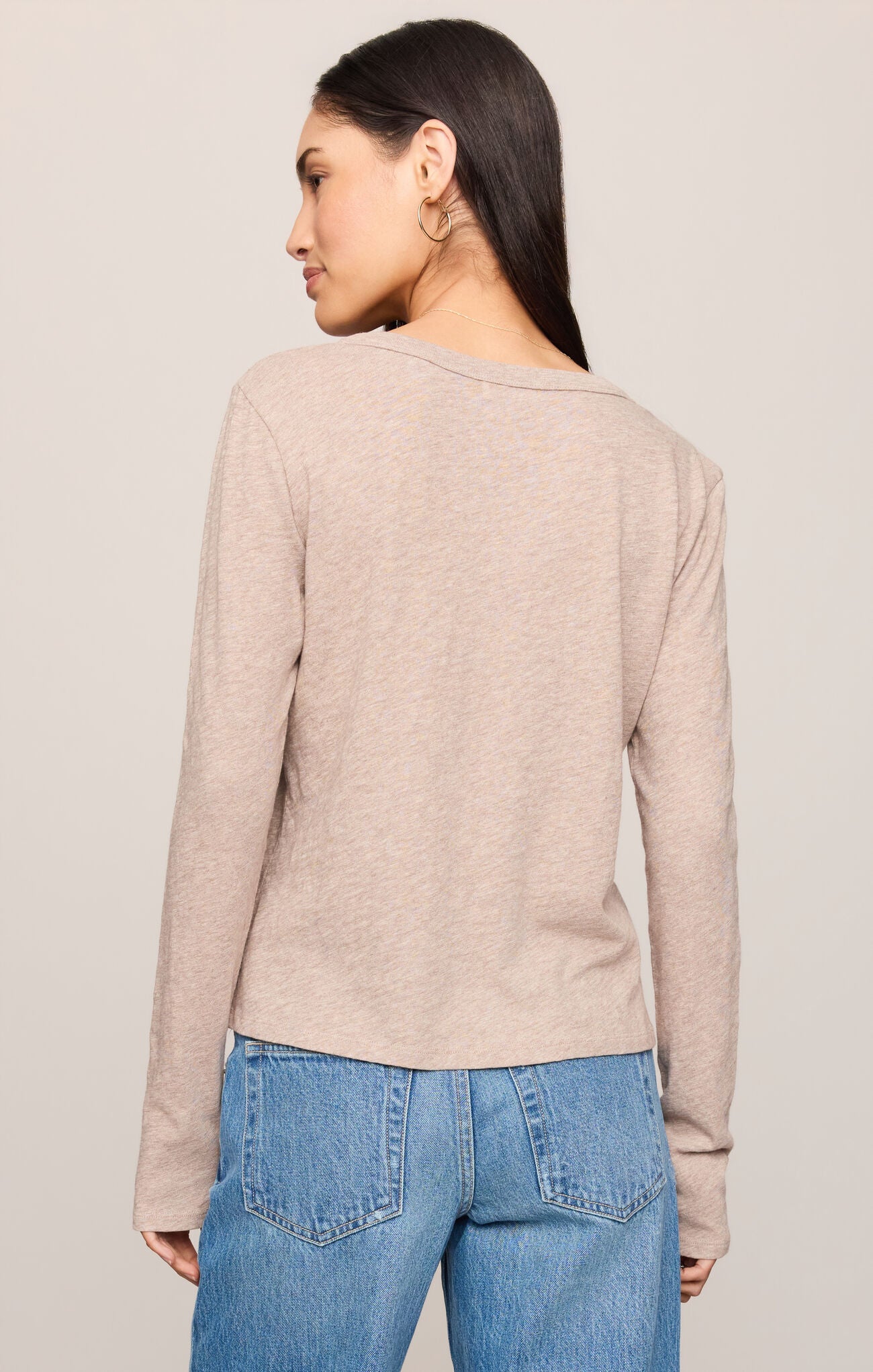 A woman shown from the back wearing a light beige long-sleeve knit top and blue jeans against a plain light gray studio background.