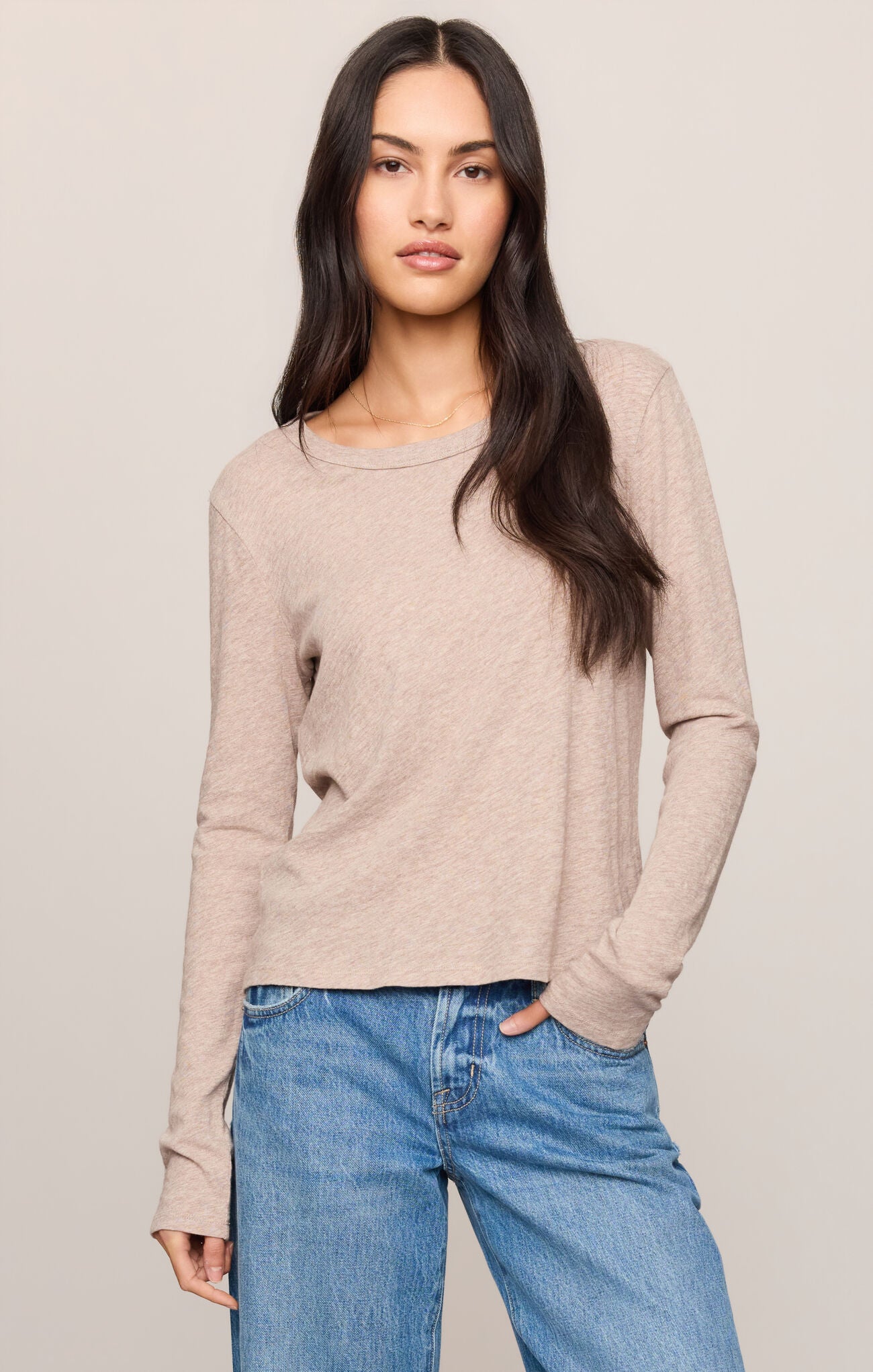 A woman modeling a light beige long-sleeve knit top with a crew neckline, paired with blue jeans, against a solid light gray background.