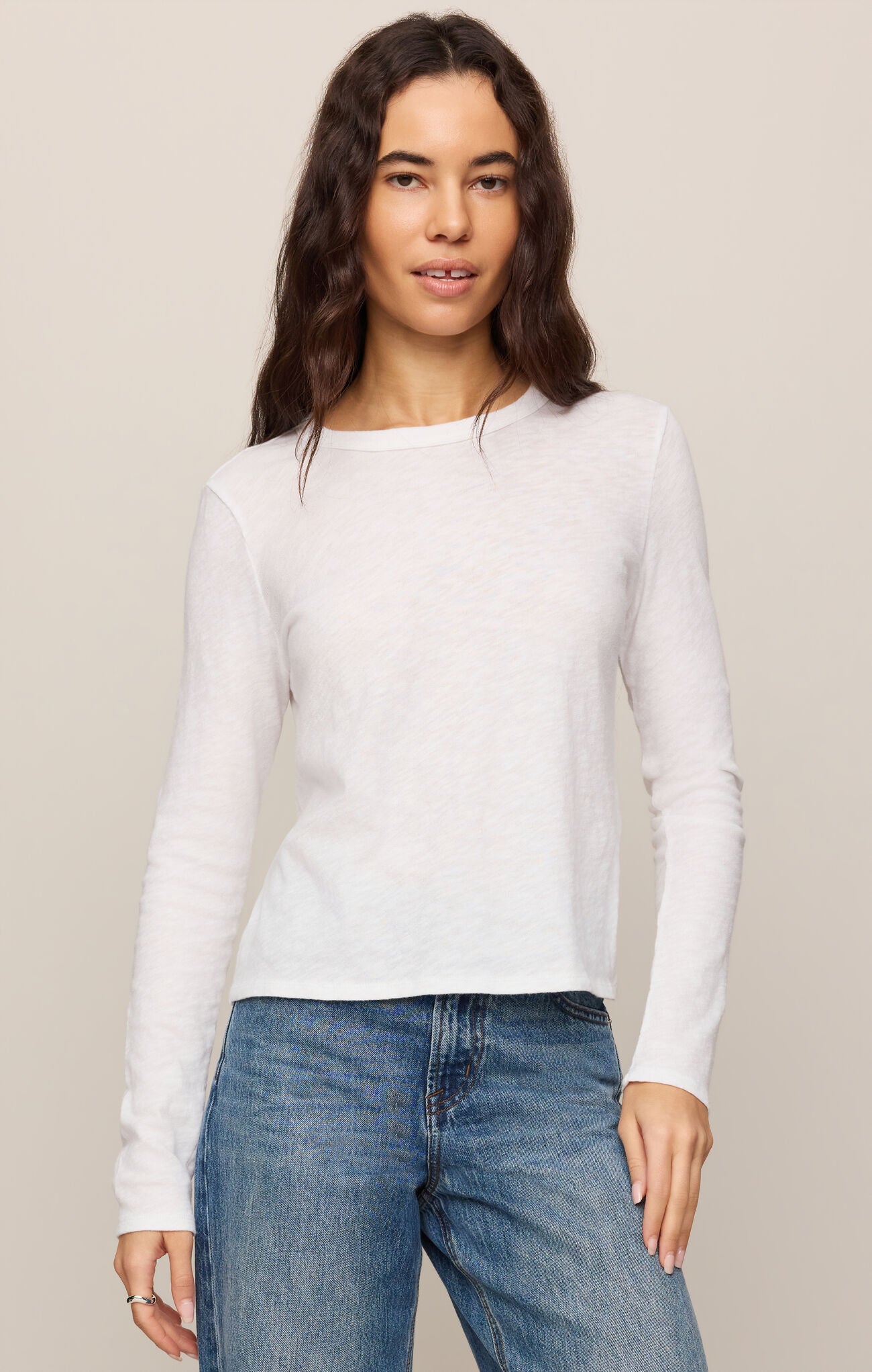 Woman wearing a white long-sleeve top and blue jeans against a beige background