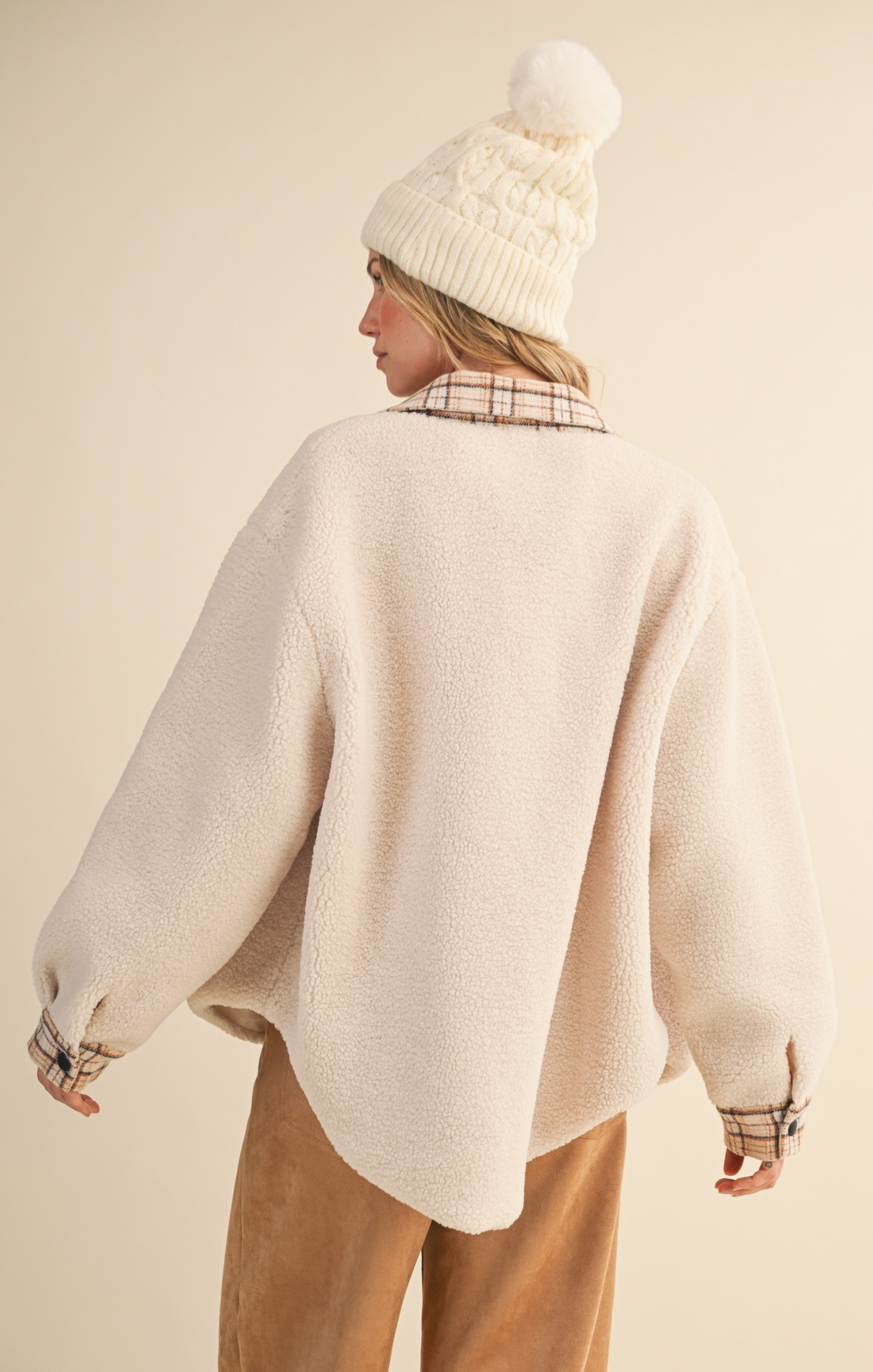 Person wearing a beige sweater and plaid shirt with a white knit hat on a beige background