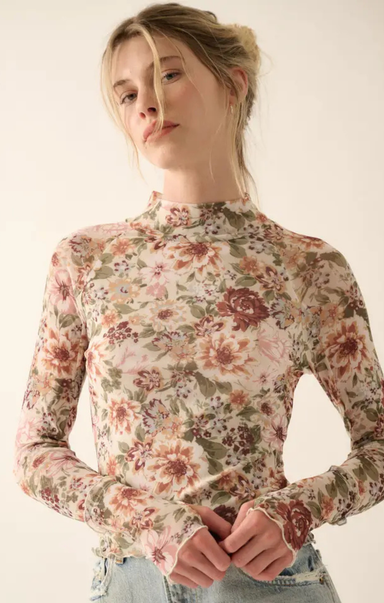 A woman wearing a long-sleeve mock neck top with a dense floral pattern in pink, red, and green on a light beige background, paired with light blue jeans.