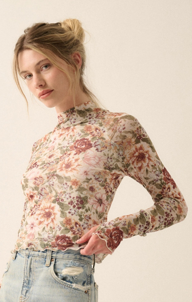 A woman wearing a long-sleeve, high-neck floral mesh top with ruffled trim, paired with light-wash distressed jeans, set against a plain off-white background.