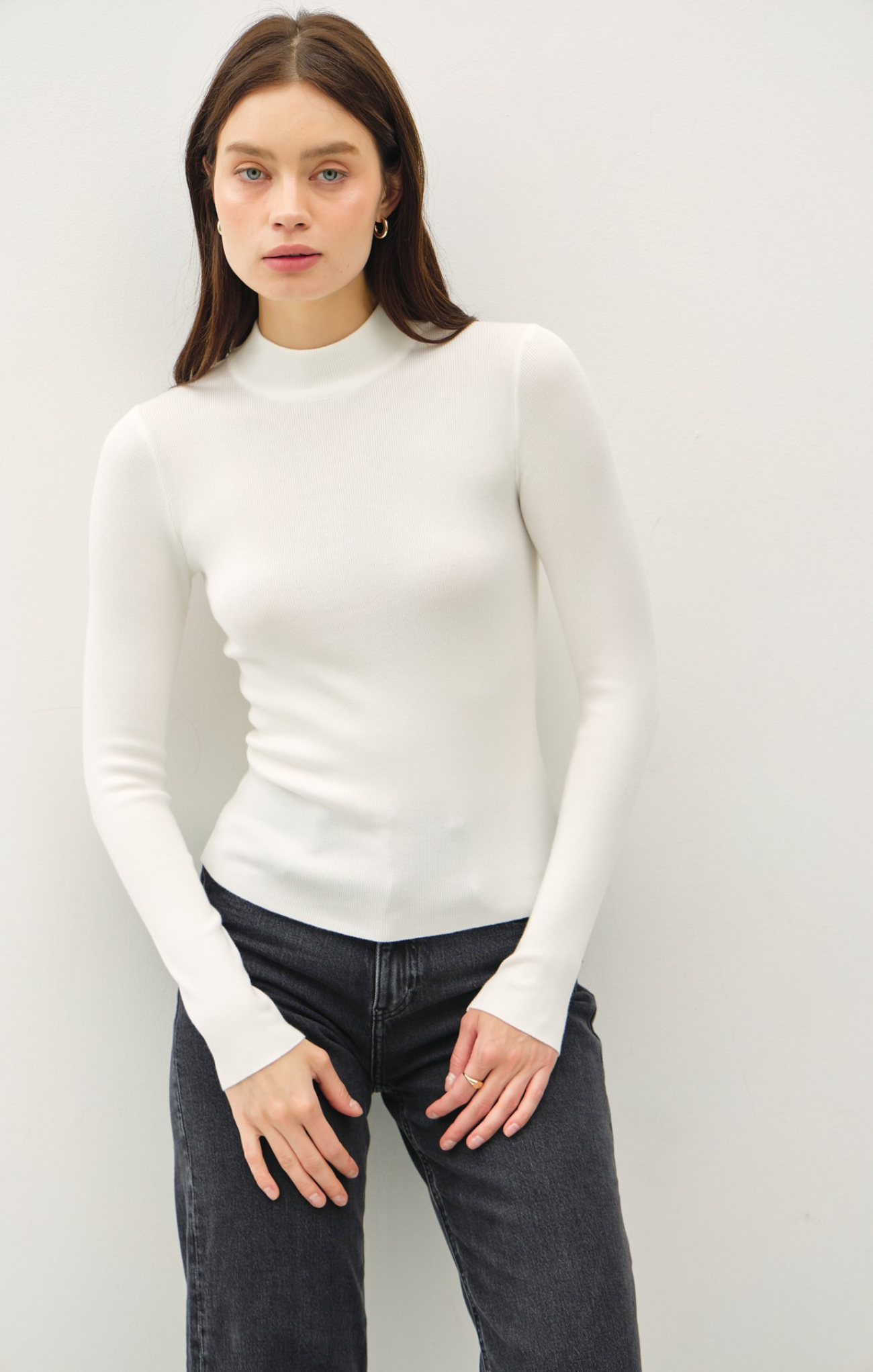 Woman wearing a white turtleneck sweater and dark jeans against a plain background