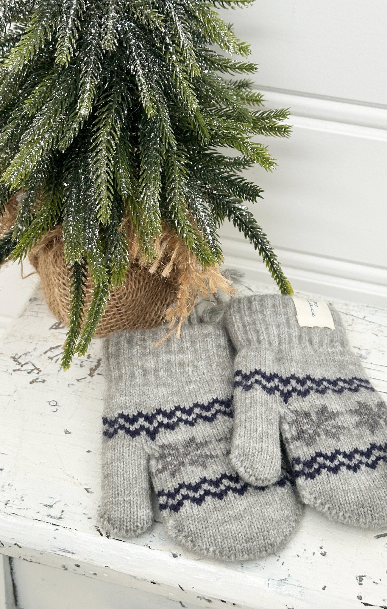 Pair of gray knitted mittens with a pattern on a white surface next to a small Christmas tree.