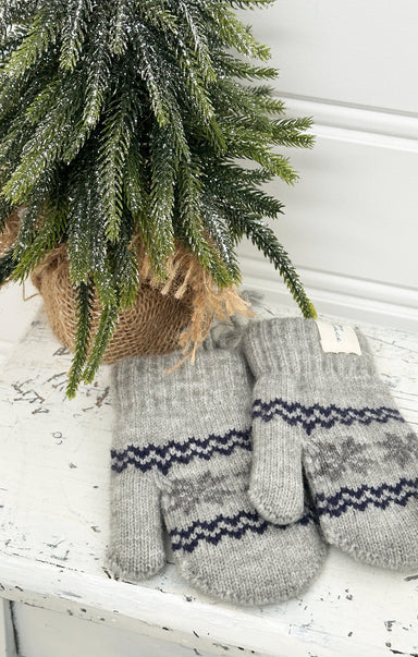 Pair of gray knitted mittens with a pattern on a white surface next to a small Christmas tree.