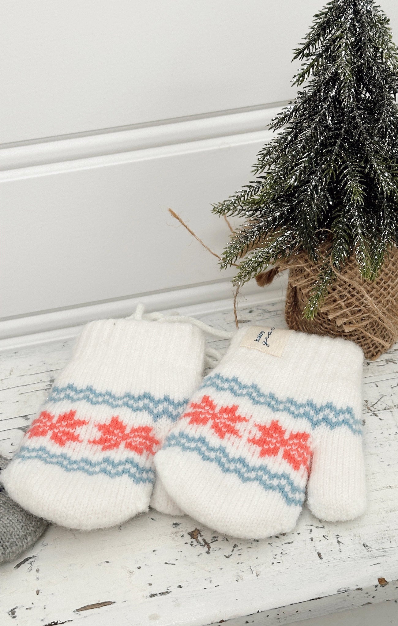Pair of white mittens with red and blue patterns on a wooden surface with a small Christmas tree in the background.
