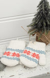 Pair of white mittens with red and blue patterns on a wooden surface with a small Christmas tree in the background.