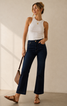 A woman modeling dark blue flare jeans with a white ribbed tank top and brown sandals, holding a brown shoulder bag against a warm beige studio background.