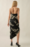 A woman shown from the back wearing a black spaghetti-strap midi dress with a beige floral print, paired with black high heels, against a plain light beige studio background.
