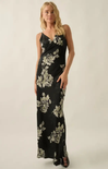 A woman modeling a sleeveless black maxi dress with a large beige floral print, photographed against a plain light beige studio background.
