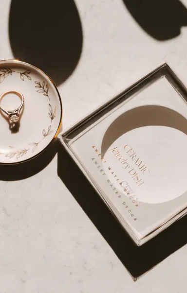 A ceramic jewelry dish with a gold ring displayed next to its square packaging box on a light marble surface with dramatic shadows.