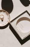 A ceramic jewelry dish with a gold ring displayed next to its square packaging box on a light marble surface with dramatic shadows.