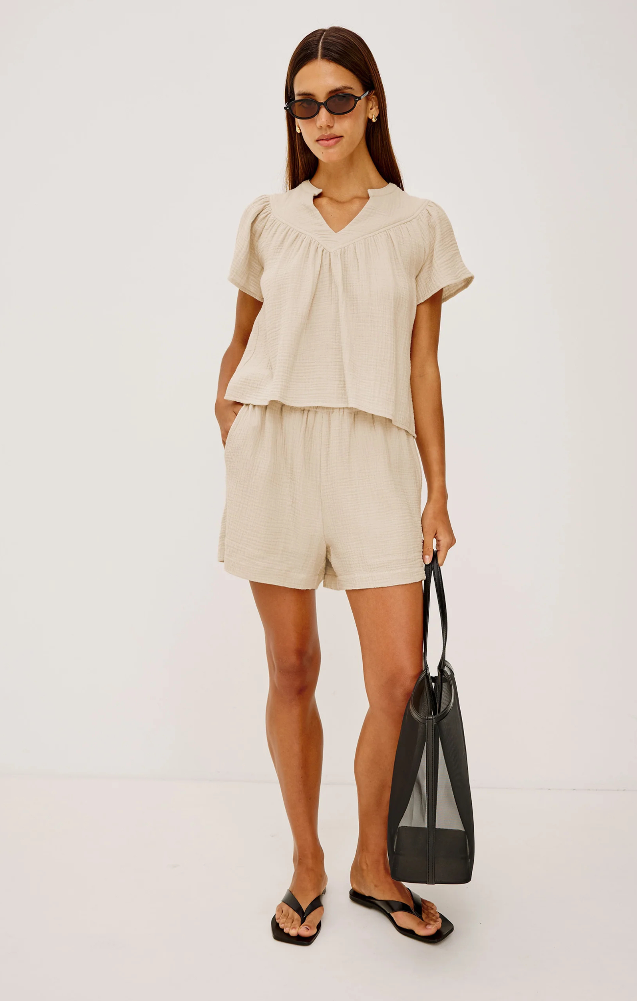 Woman wearing a beige outfit with shorts and a top, holding a black bag on a white background