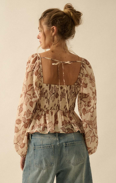 Woman wearing a floral blouse with a sheer back and blue jeans on a beige background