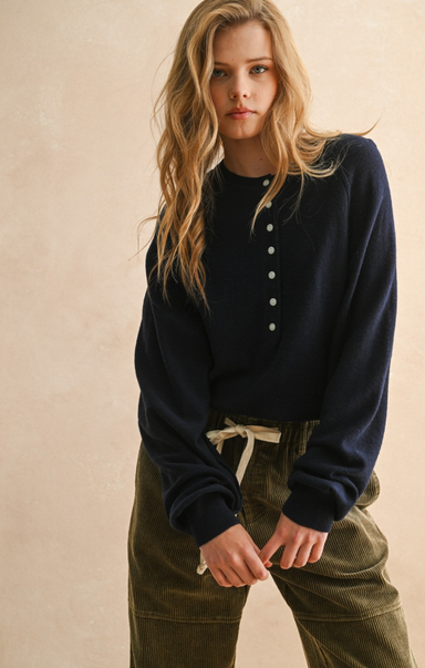 Woman wearing a navy sweater and green pants against a beige background