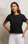 A woman modeling a black short-sleeve crewneck t-shirt paired with white wide-leg pants, standing against a light beige fabric backdrop.