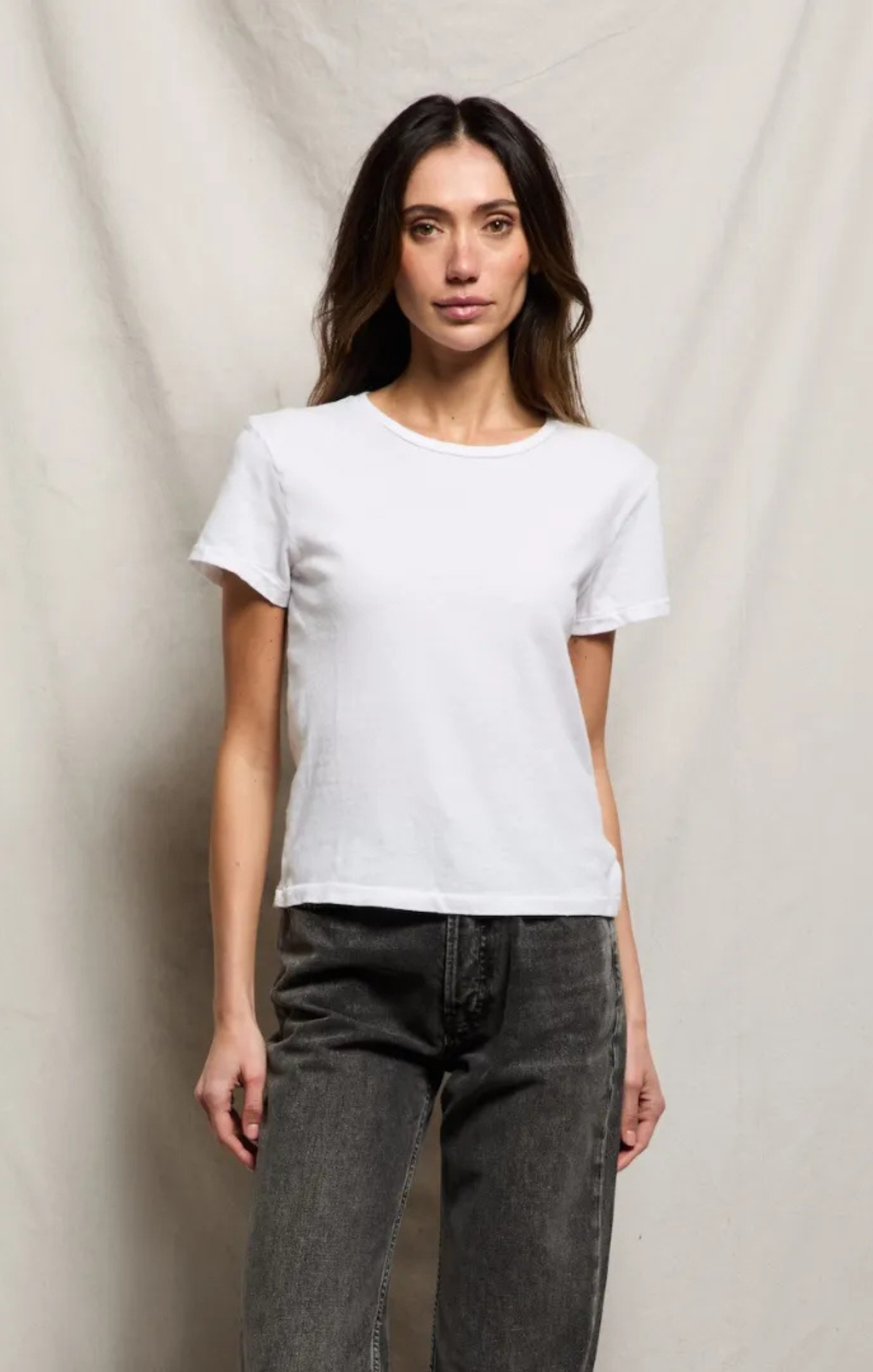 A woman modeling a plain white short-sleeve crewneck t-shirt paired with dark gray jeans, standing against a draped beige fabric backdrop.