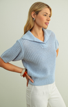 Woman wearing a light blue knitted sweater and white pants against a plain background