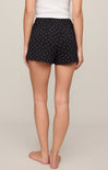 A person shown from the back wearing black shorts with a small white polka dot pattern, paired with a white top, against a plain light beige studio background.