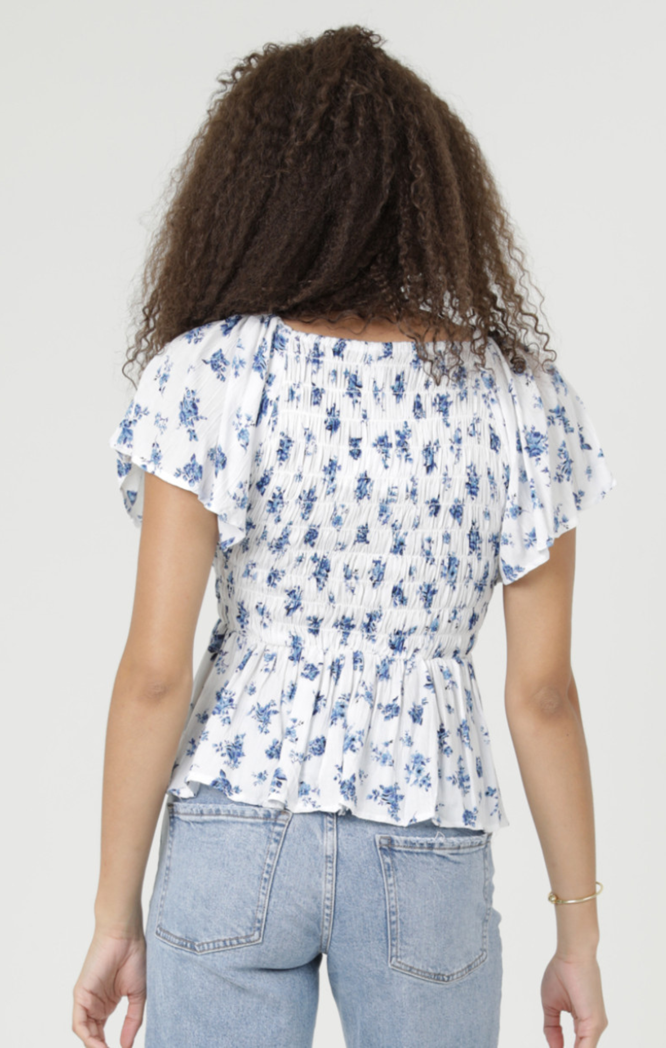 Person wearing a white blouse with blue floral pattern and light blue jeans on a plain background