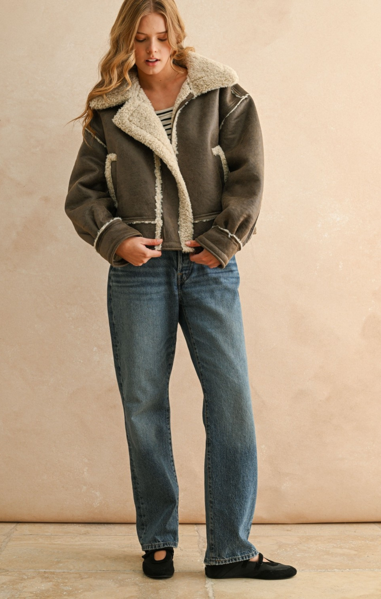 Woman wearing a brown shearling jacket and blue jeans against a beige background
