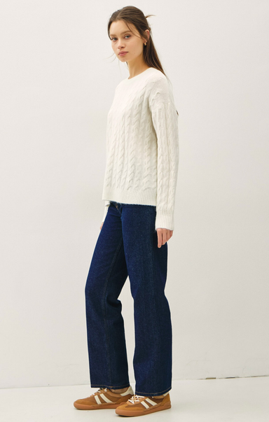 Woman wearing a white cable knit sweater and dark blue jeans on a white background