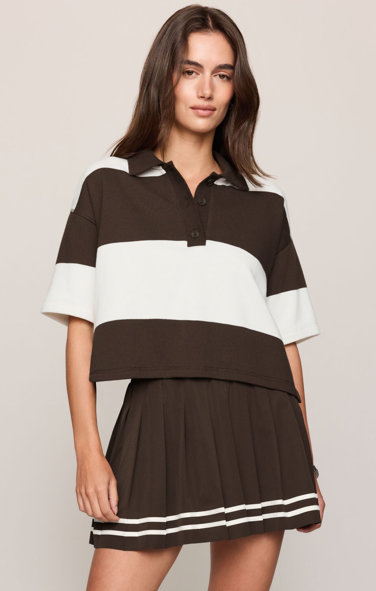 A woman modeling a brown and white color-blocked short-sleeve polo shirt with a matching pleated mini skirt against a light beige studio background.