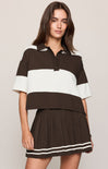 A woman modeling a brown and white color-blocked short-sleeve polo shirt with a matching pleated mini skirt against a light beige studio background.