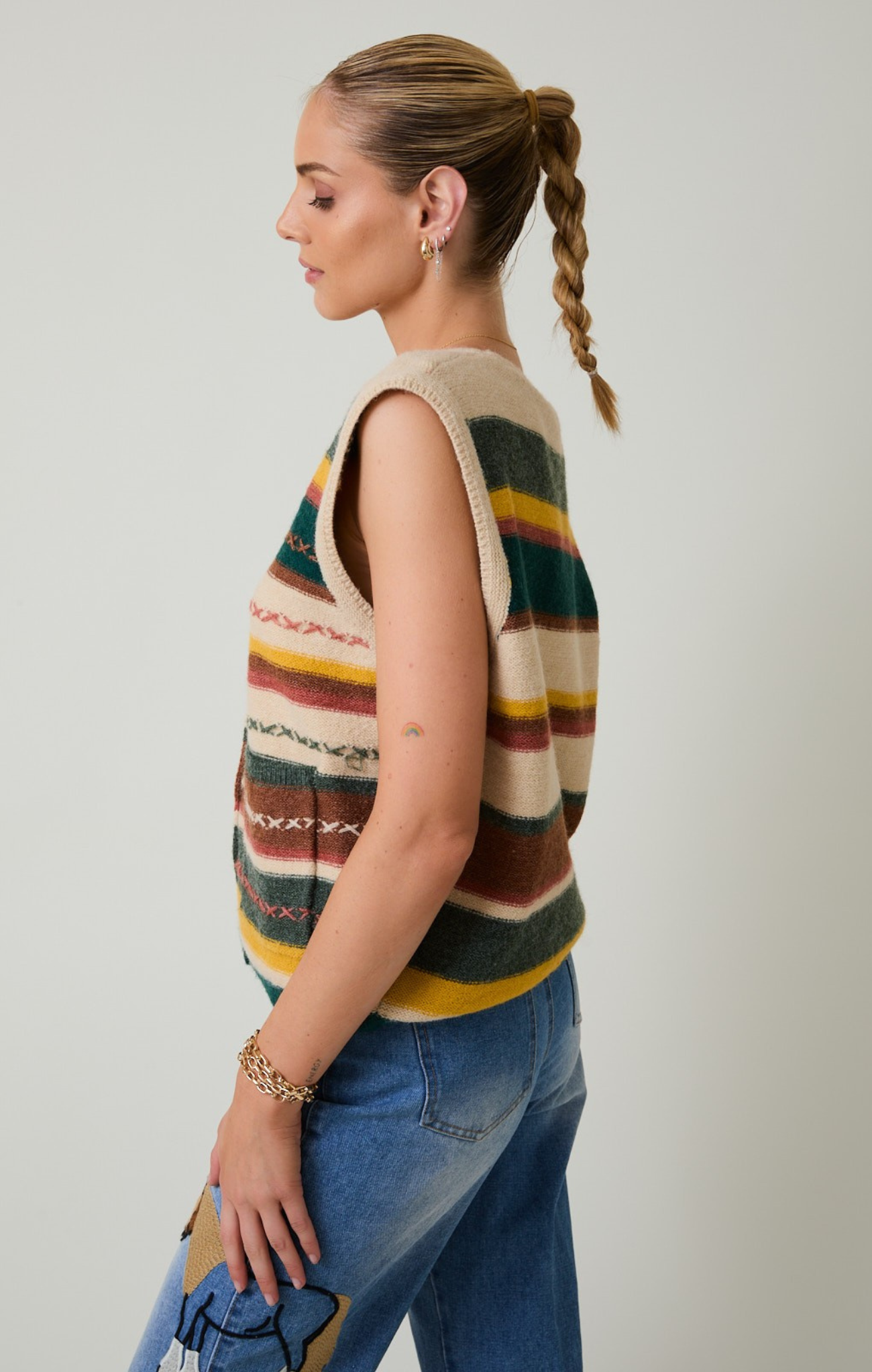 Woman wearing a colorful striped sleeveless sweater and blue jeans against a plain background
