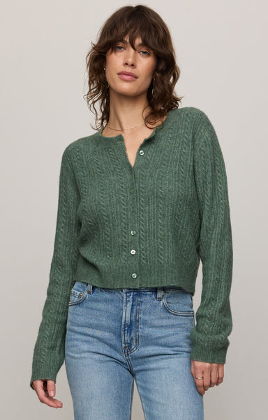 Woman wearing a green knitted cardigan and blue jeans against a plain background