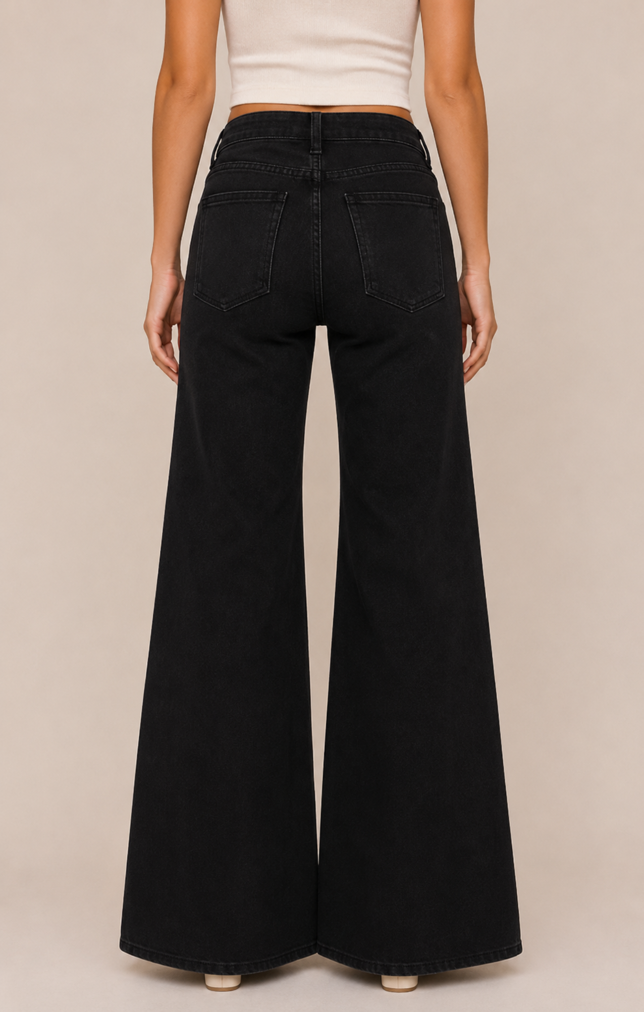 Rear view of a person wearing black wide-leg jeans with a white cropped top, standing against a plain beige studio background.