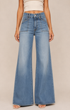 A model wearing high-waisted, wide-leg blue denim jeans with a light wash, styled with a cream ribbed crop top and clear high-heel sandals against a solid beige studio background.