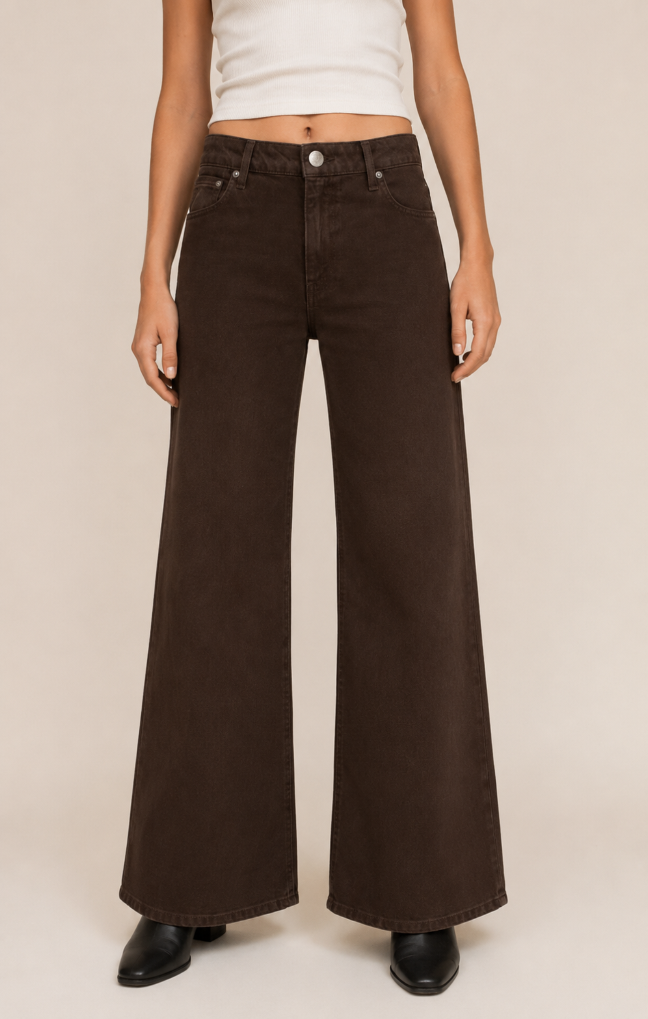 A model wearing dark brown wide-leg denim jeans, a white cropped tank top, and black shoes, standing against a plain beige studio background.