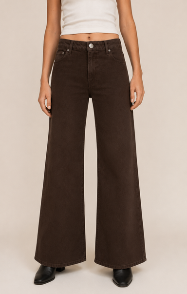 A model wearing dark brown wide-leg denim jeans, a white cropped tank top, and black shoes, standing against a plain beige studio background.