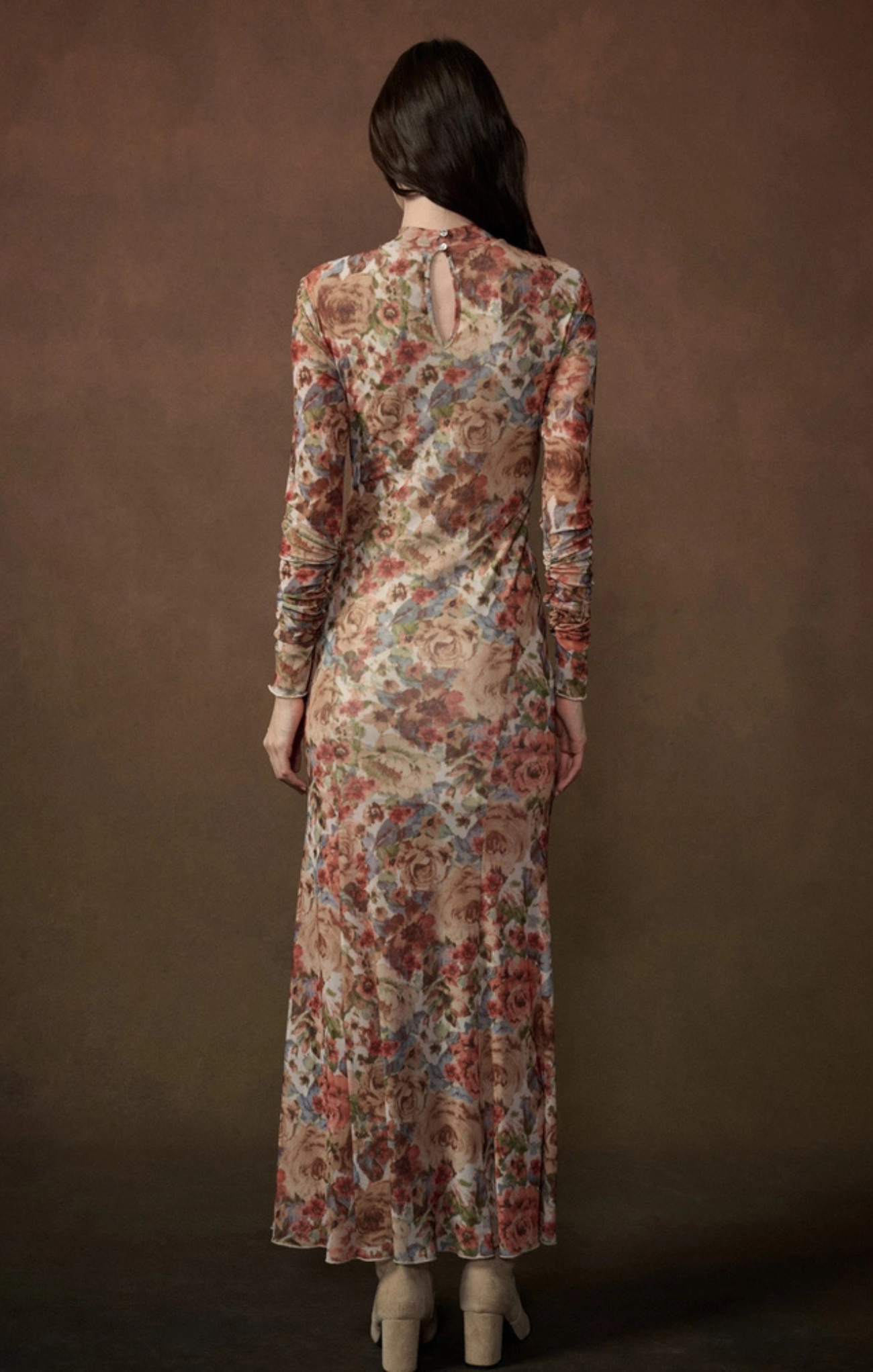 A woman viewed from the back wearing a long-sleeve maxi dress with a dense floral pattern in muted red, blue, and beige tones, set against a solid dark brown studio background.