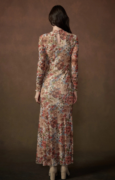A woman viewed from the back wearing a long-sleeve maxi dress with a dense floral pattern in muted red, blue, and beige tones, set against a solid dark brown studio background.
